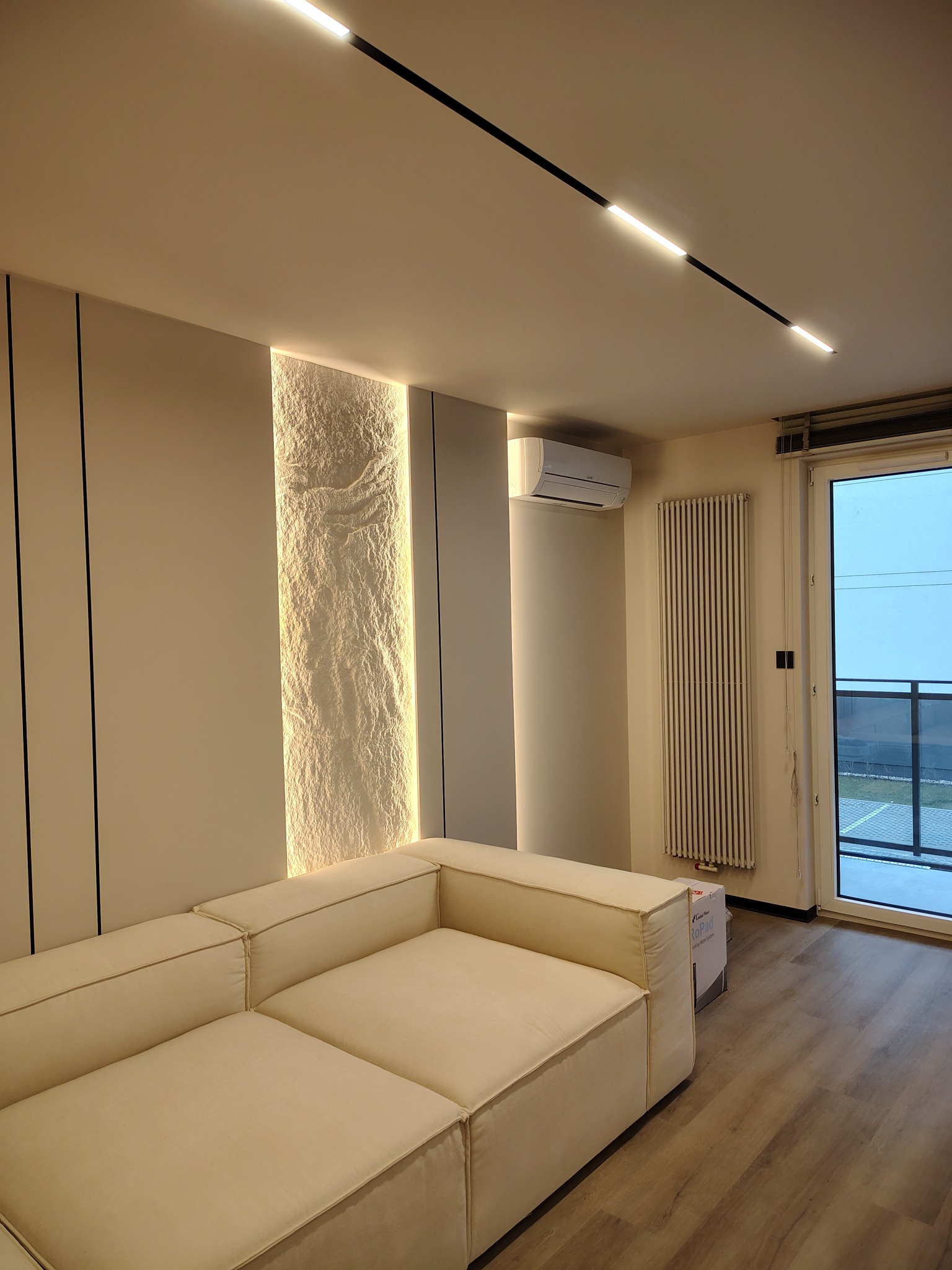 Nowoczesny salon z beżową sofą, podświetlaną ścianą z efektem kamienia i minimalistycznym oświetleniem sufitowym. Widoczny grzejnik i klimatyzator.