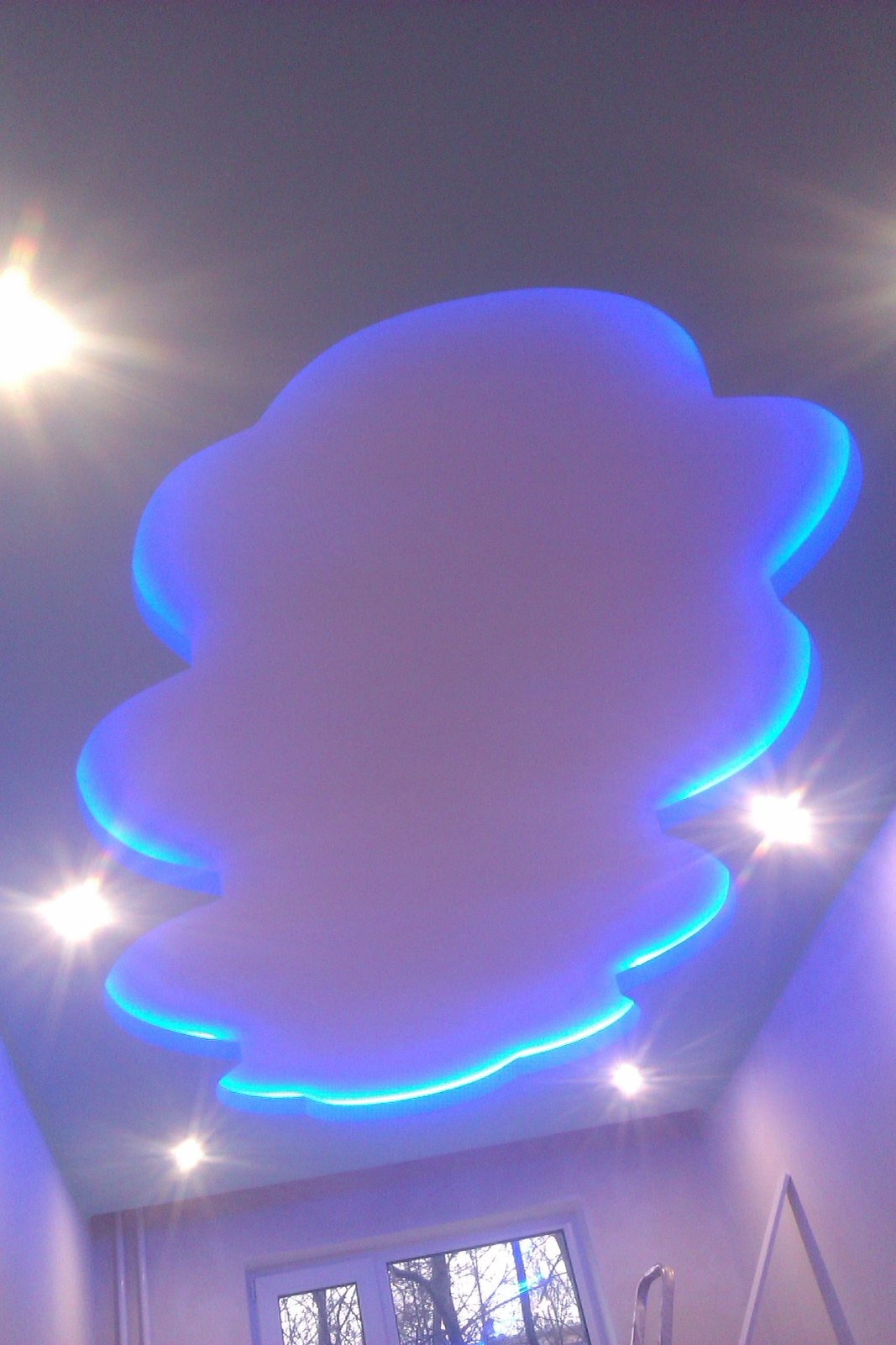 Podwieszany sufit w kształcie chmury z niebieskim podświetleniem LED, widok z dołu, okno w tle. Nietypowy design sufitu w pokoju.