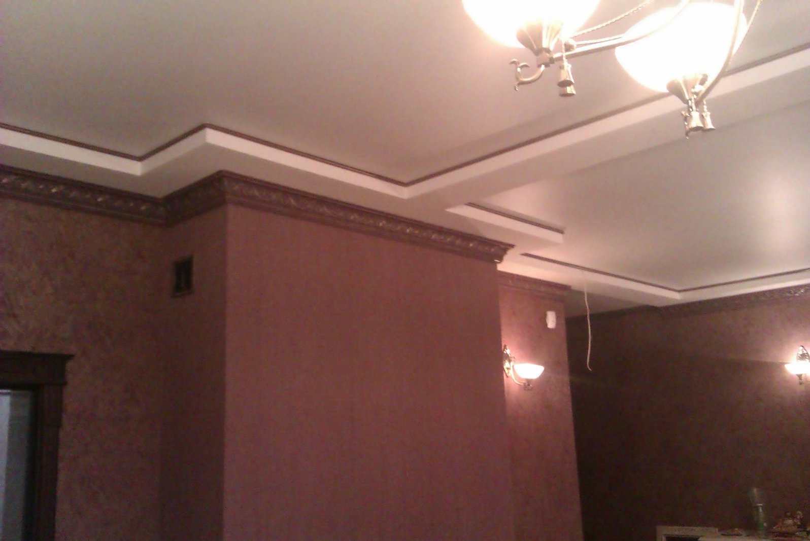 Eleganckie wykończenie wnętrza z dekoracyjną sztukaterią sufitową i bordowymi ścianami. Widoczne oświetlenie ścienne i sufitowe.