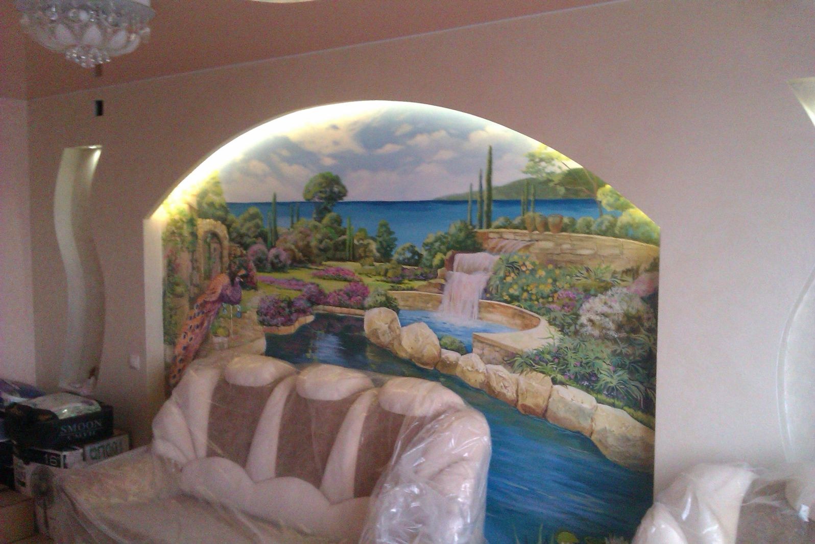 Malowidło ścienne w łuku przedstawiające pejzaż z wodospadem, roślinnością i pawiem, częściowo zasłonięte przez meble zabezpieczone folią.