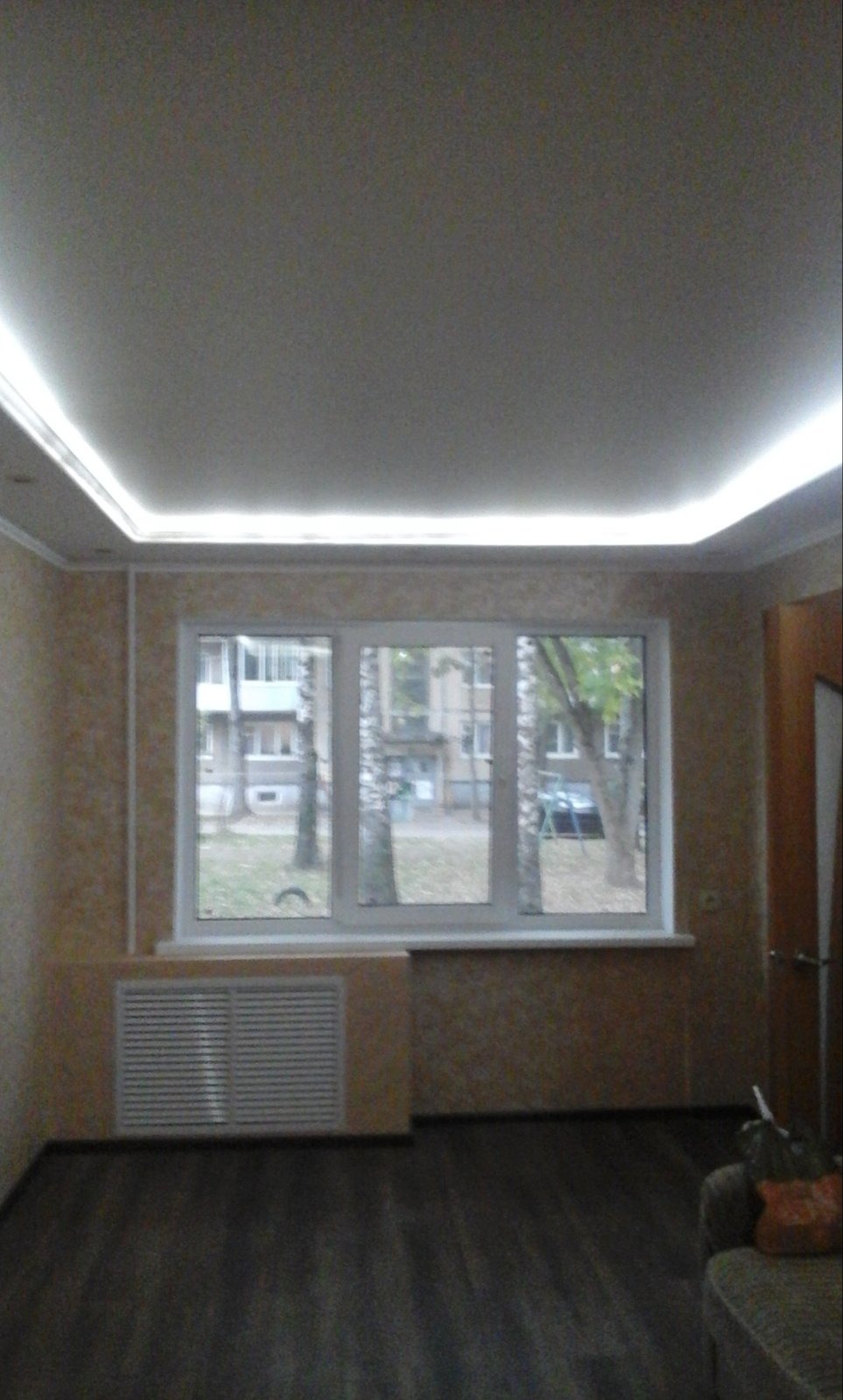 Jasne wnętrze pokoju po remoncie: kremowe ściany z fakturą, okno z widokiem na drzewa, ciemna podłoga i sufit z oświetleniem LED.