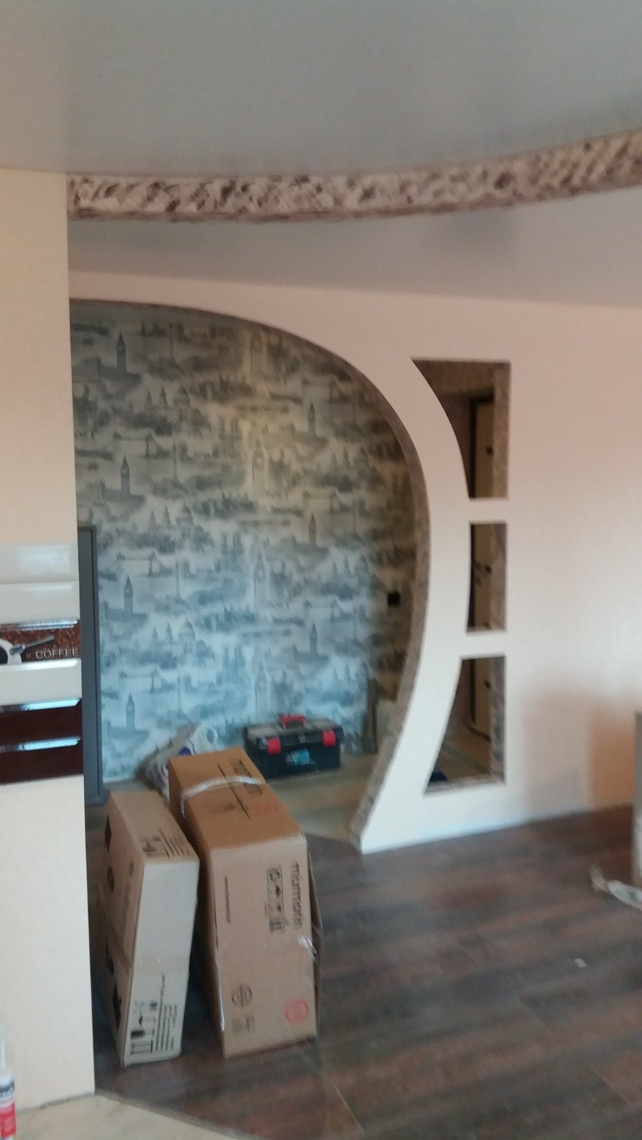 Aranżacja wnętrza z dekoracyjnym łukiem i tapetą w stylu miejskim. Widoczne kartony i narzędzia, sugerujące etap prac wykończeniowych.