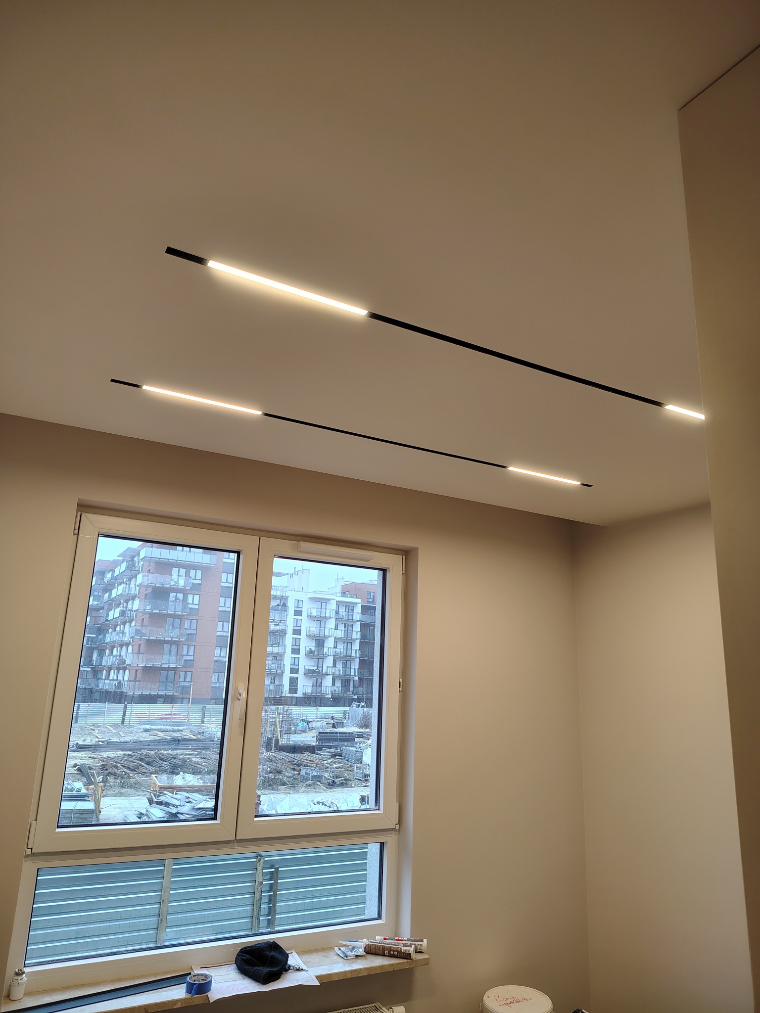 Minimalistyczne oświetlenie sufitowe z czarnymi listwami LED w jasnym pokoju z oknem na budowę. Widok na budynki w tle.