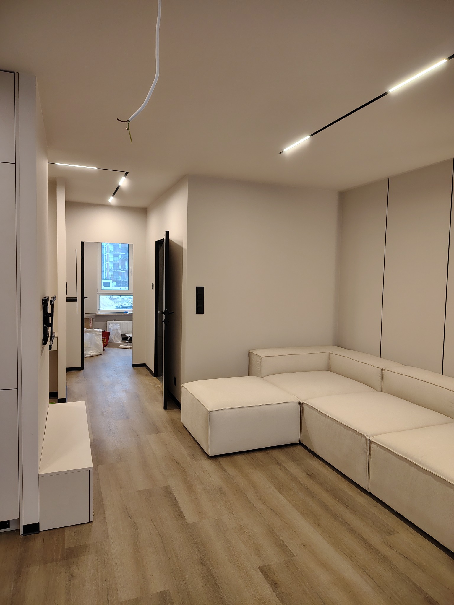 Jasne wnętrze po remoncie: narożna sofa, panele podłogowe, minimalistyczne oświetlenie sufitowe. W tle widoczne materiały budowlane, sugerujące świeżo zakończone prace.