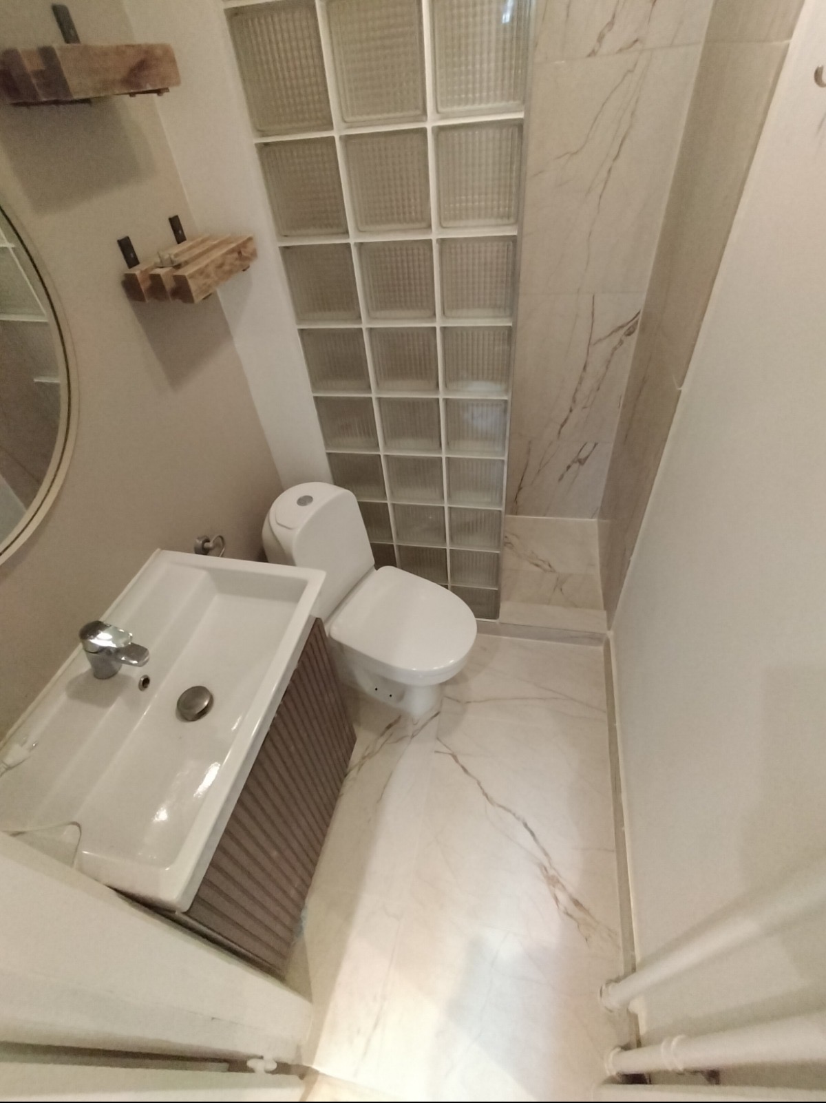 Jasna łazienka po remoncie: biała umywalka z szafką, toaleta, marmurowe płytki podłogowe i ścienne, ściana z luksferów. Drewniane półki nad umywalką.