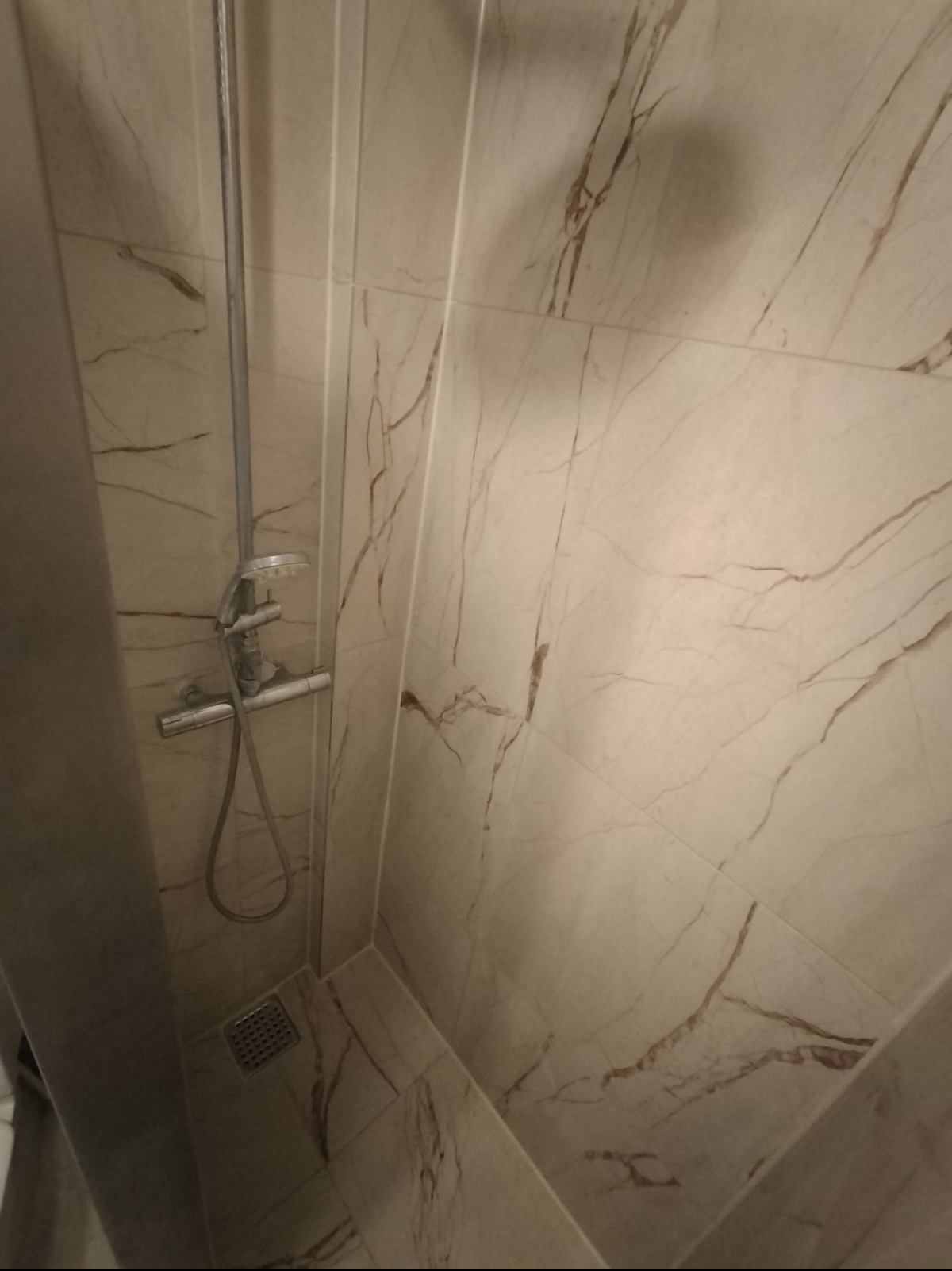 Wykończona kabina prysznicowa z beżowymi płytkami imitującymi marmur. Widoczna bateria prysznicowa i odpływ liniowy. Minimalistyczny design, ciepła kolorystyka.