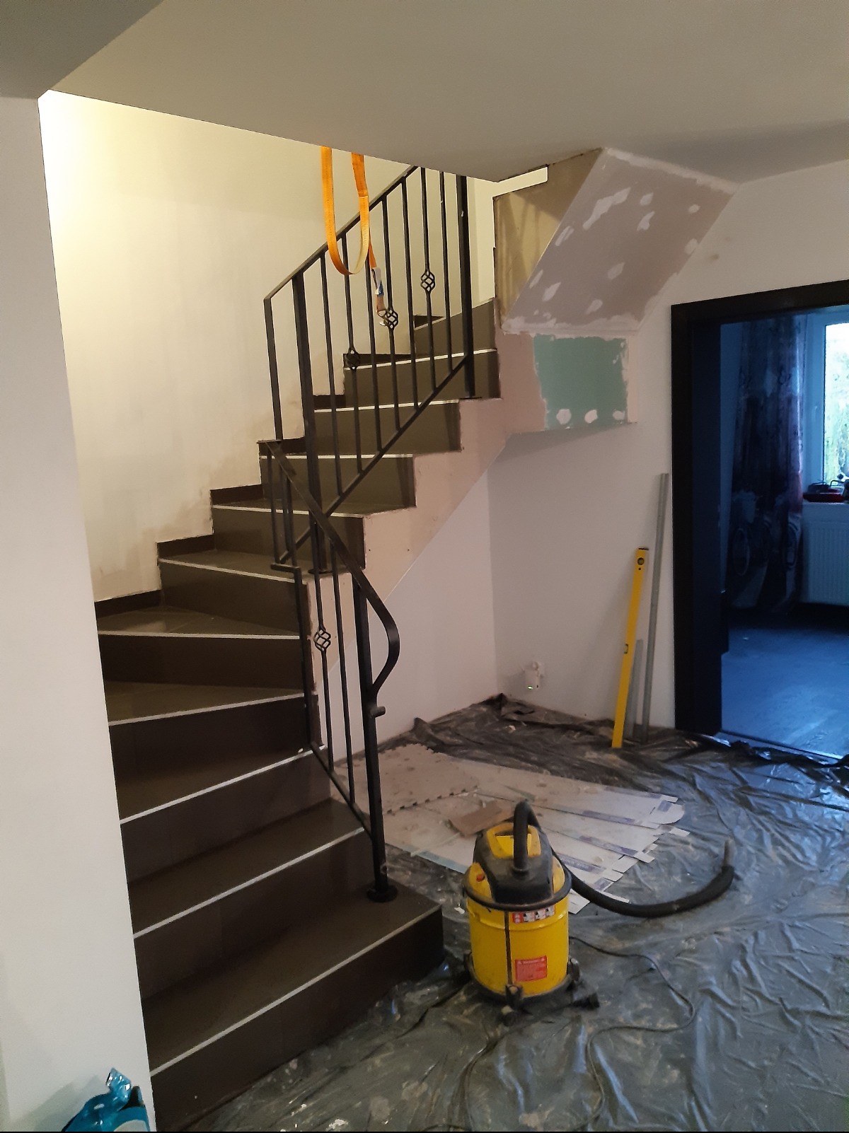 Klatka schodowa w trakcie remontu: brązowe stopnie, czarna balustrada, żółty odkurzacz przemysłowy, folia ochronna na podłodze, fragment ściany z gładzią.