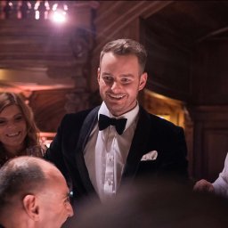 Just Edi Show - Uśmiechnięty mężczyzna w smokingu, otoczony gośćmi podczas eleganckiego przyjęcia. W tle dekoracyjne elementy architektoniczne i oświetlenie.