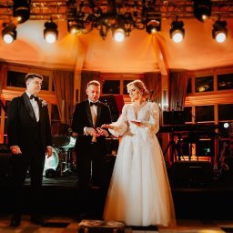 Just Edi Show - Iluzjonista prezentuje sztuczkę z kartami na scenie podczas wesela. W tle zespół muzyczny i oświetlenie sceniczne. Para młoda obserwuje z zaciekawieniem.