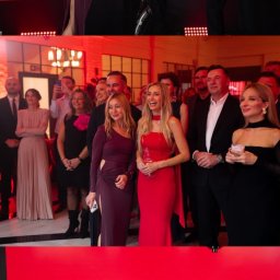 Just Edi Show - Iluzjonista prezentuje triki z kartami na scenie podczas gali. W tle widownia i czerwone tło z logo. Mężczyzna w smokingu i kobiety w eleganckich sukniach.