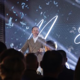 Just Edi Show - Mężczyzna w brązowym garniturze prezentuje pokaz iluzji na scenie przed publicznością, z podświetlonym tłem i mikrofonem nagłownym.