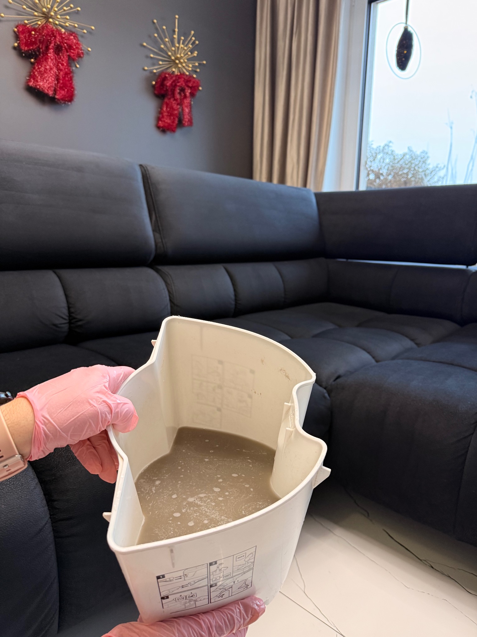Brudna woda po praniu tapicerki narożnika w Kluczborku, widoczna w białym pojemniku trzymanym w rękawicach ochronnych. W tle ciemny narożnik i dekoracje.