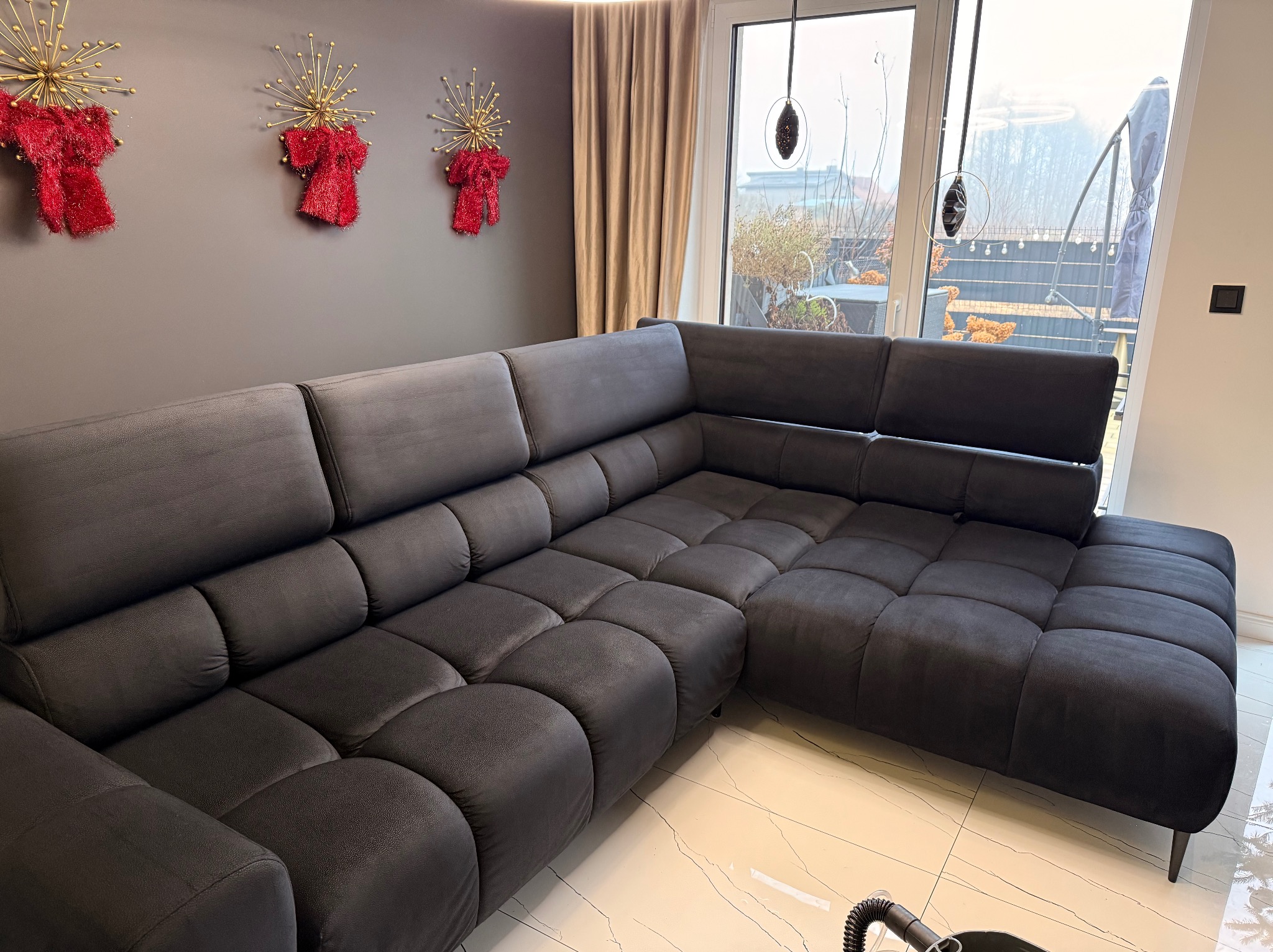 Ciemnoszara, modułowa kanapa narożna z weluru w nowoczesnym salonie z dekoracjami ściennymi i jasną podłogą. Widoczny fragment odkurzacza.