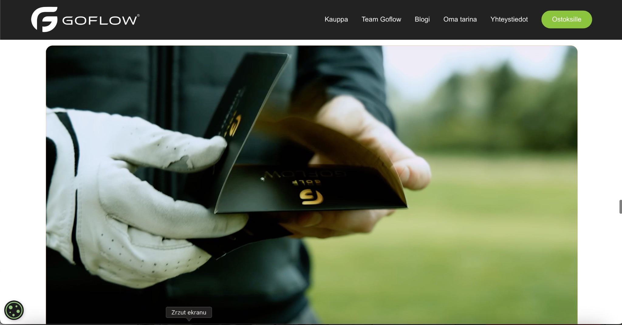 Dłoń w białej rękawicy golfowej prezentuje czarne opakowanie z logo GoFlow Golf na tle zielonego pola golfowego. Opakowanie ma złote akcenty.