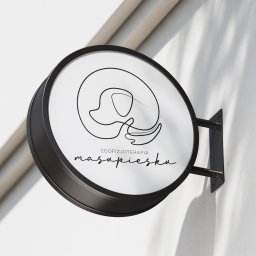 Katarzyna Rębacz - Okrągły szyld z minimalistycznym logo przedstawiającym psa i napisem 'zoofizjoterapia masupiesku', zamontowany na białej ścianie w cieniu.