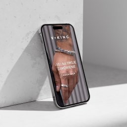 Katarzyna Rębacz - Smartfon oparty o ścianę prezentuje reklamę biżuterii V+KING z rabatem -15% na zamówienie. Na ekranie dłoń z pierścionkiem i bransoletką na tle minimalistycznym.