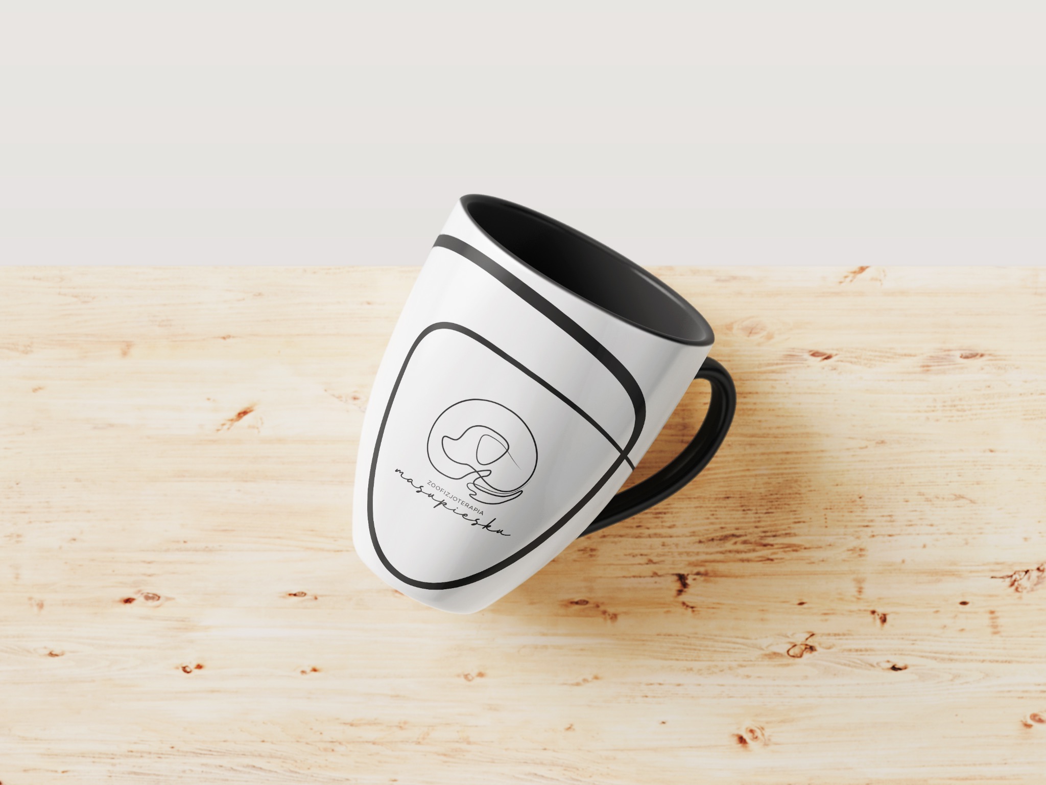 Biały kubek z czarnym wnętrzem i uchwytem, leżący na drewnianym blacie, z minimalistycznym logo i napisem 'zoofizjoterapia masapiaska'.