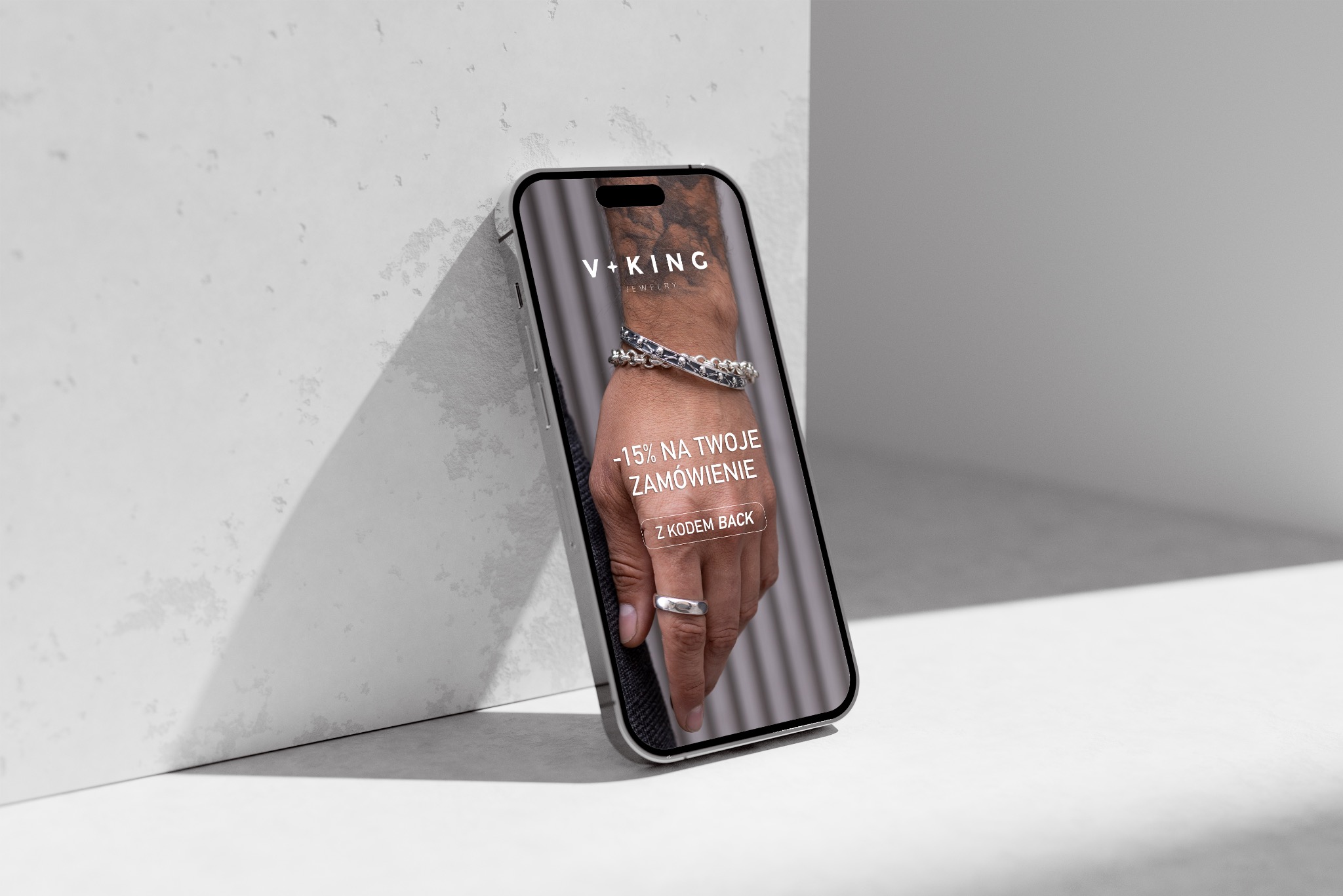 Smartfon oparty o ścianę prezentuje reklamę biżuterii V+KING z rabatem -15% na zamówienie. Na ekranie dłoń z pierścionkiem i bransoletką na tle minimalistycznym.