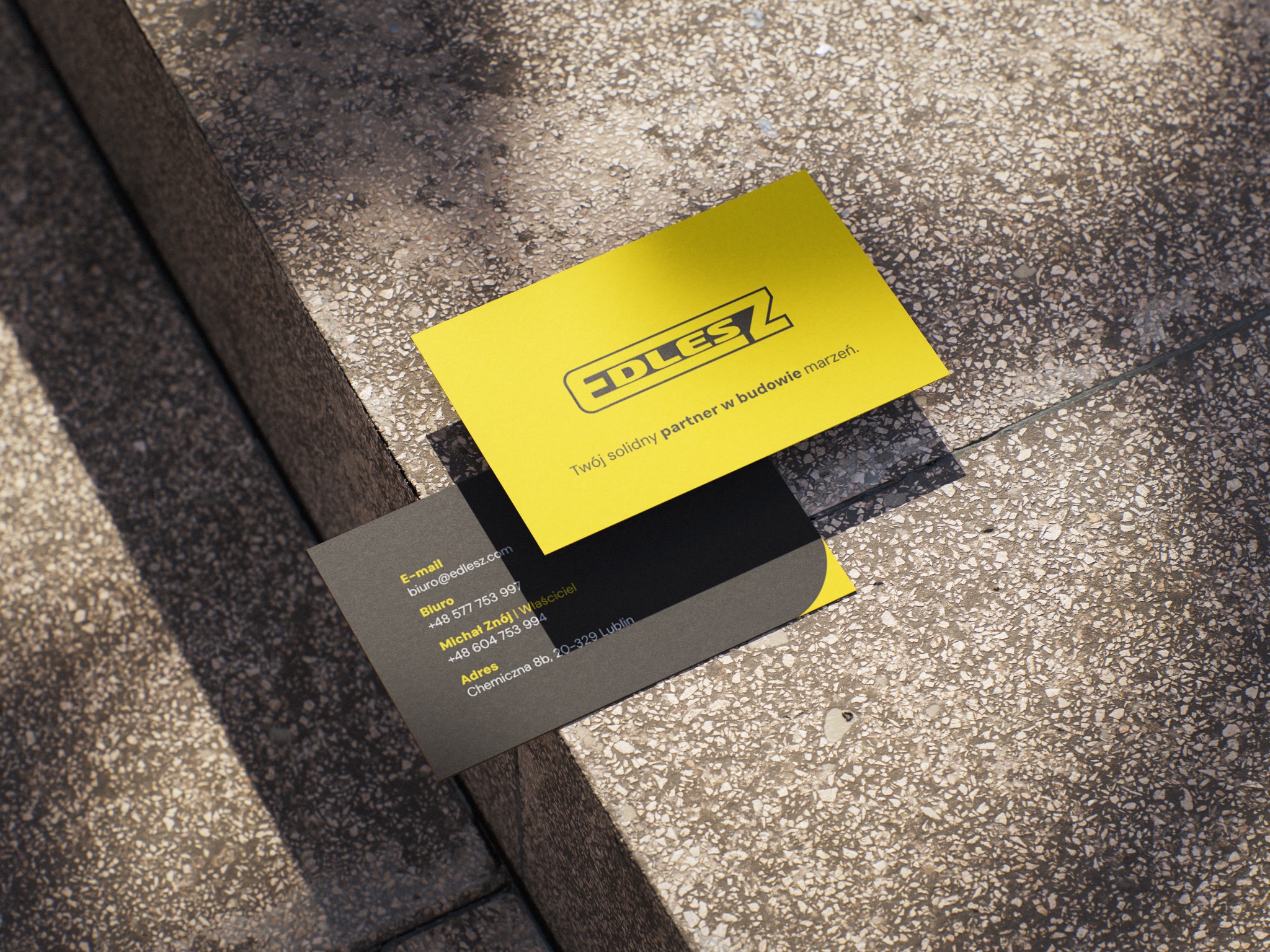 Dwie wizytówki firmy budowlanej EDLESZ leżące na betonowych schodach w ostrym świetle słonecznym. Jedna żółta z logo, druga szara z danymi kontaktowymi.