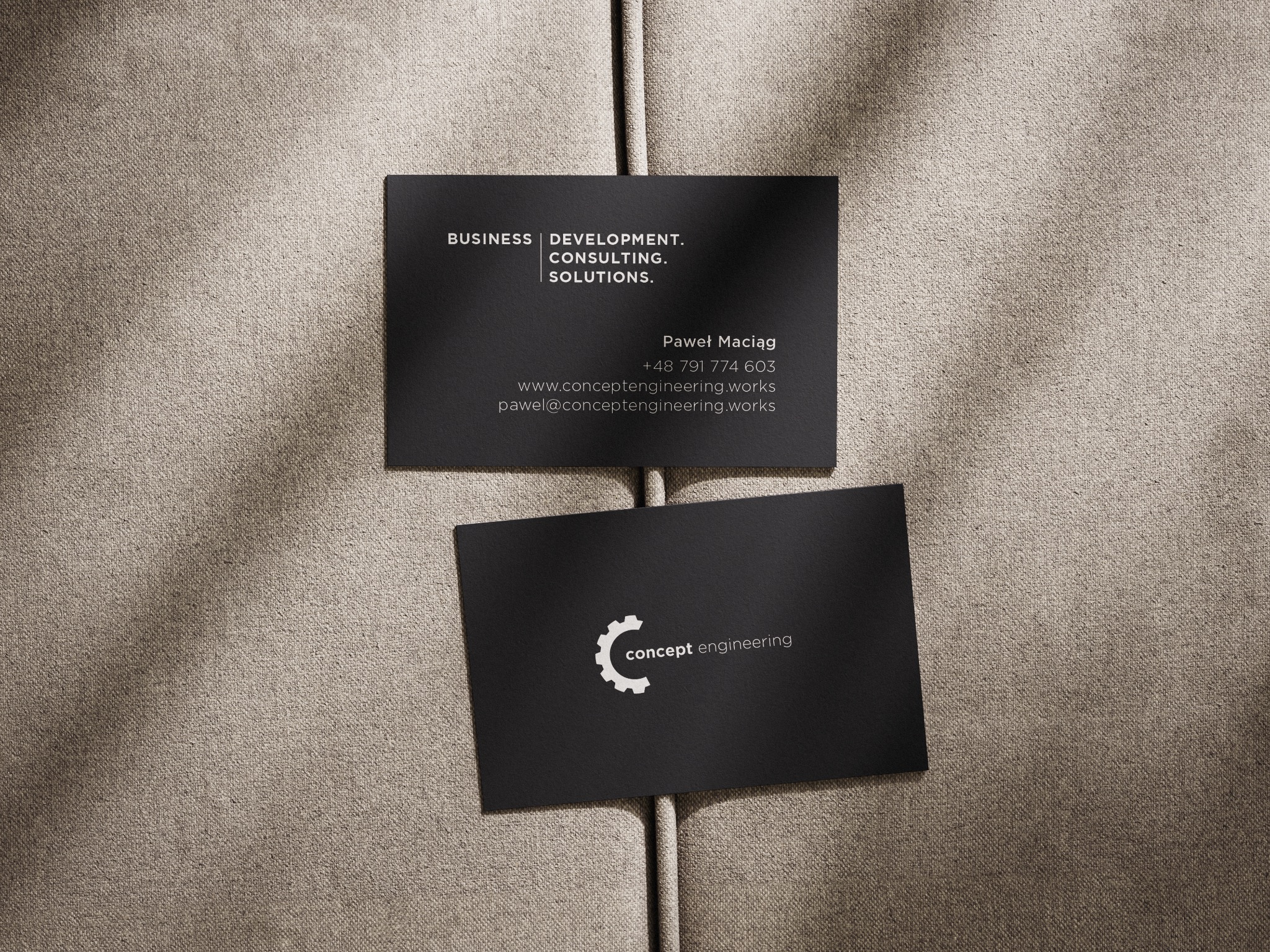 Dwie czarne wizytówki z białym tekstem i logo na teksturowanym, beżowym tle. Górna wizytówka zawiera dane kontaktowe, dolna logo 'concept engineering'.
