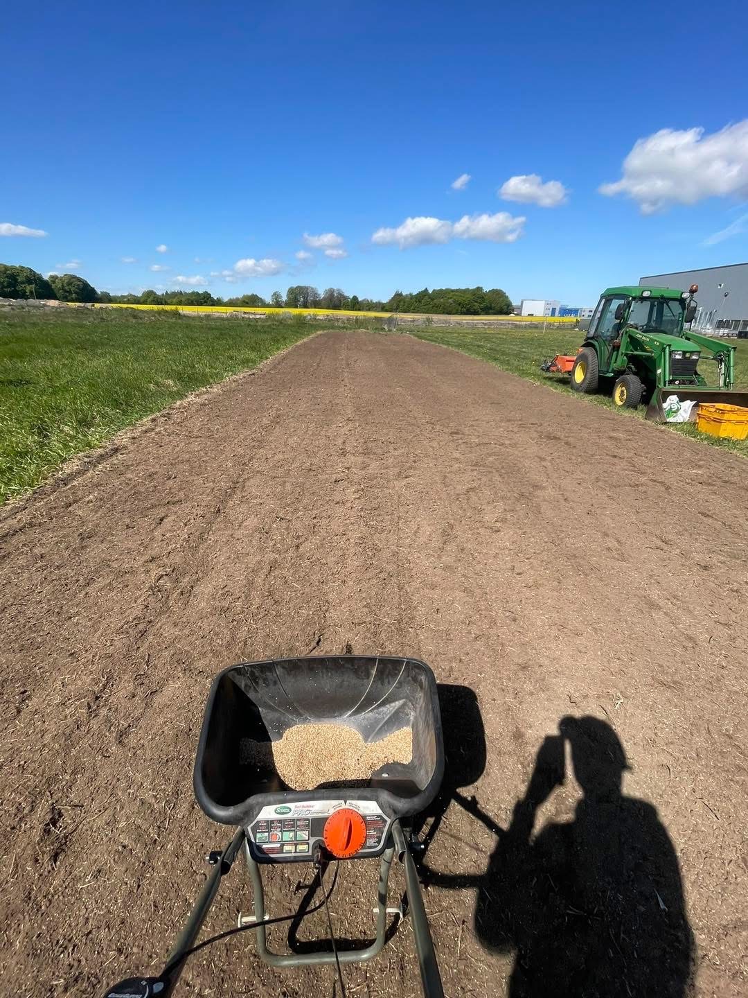 Rozsiewacz do trawy na przygotowanym terenie w Koszalinie. W tle zielony traktor i pole. Widoczne ziarna trawy w rozsiewaczu i cień osoby robiącej zdjęcie.