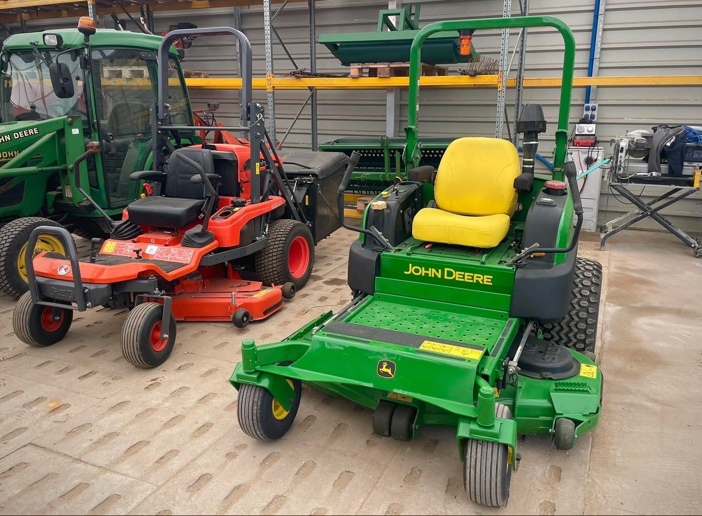 Dwie kosiarki samojezdne: zielona John Deere z żółtym siedzeniem i czerwona Kubota, stojące obok siebie w magazynie z półkami w tle. Widoczny również fragment zielonego traktora.