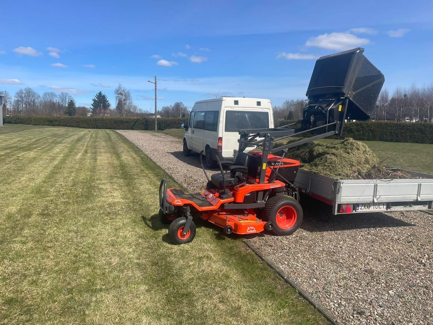 Pomarańczowa kosiarka samojezdna Kubota z przyczepą pełną skoszonej trawy na tle świeżo skoszonego trawnika i białego busa w słoneczny dzień.