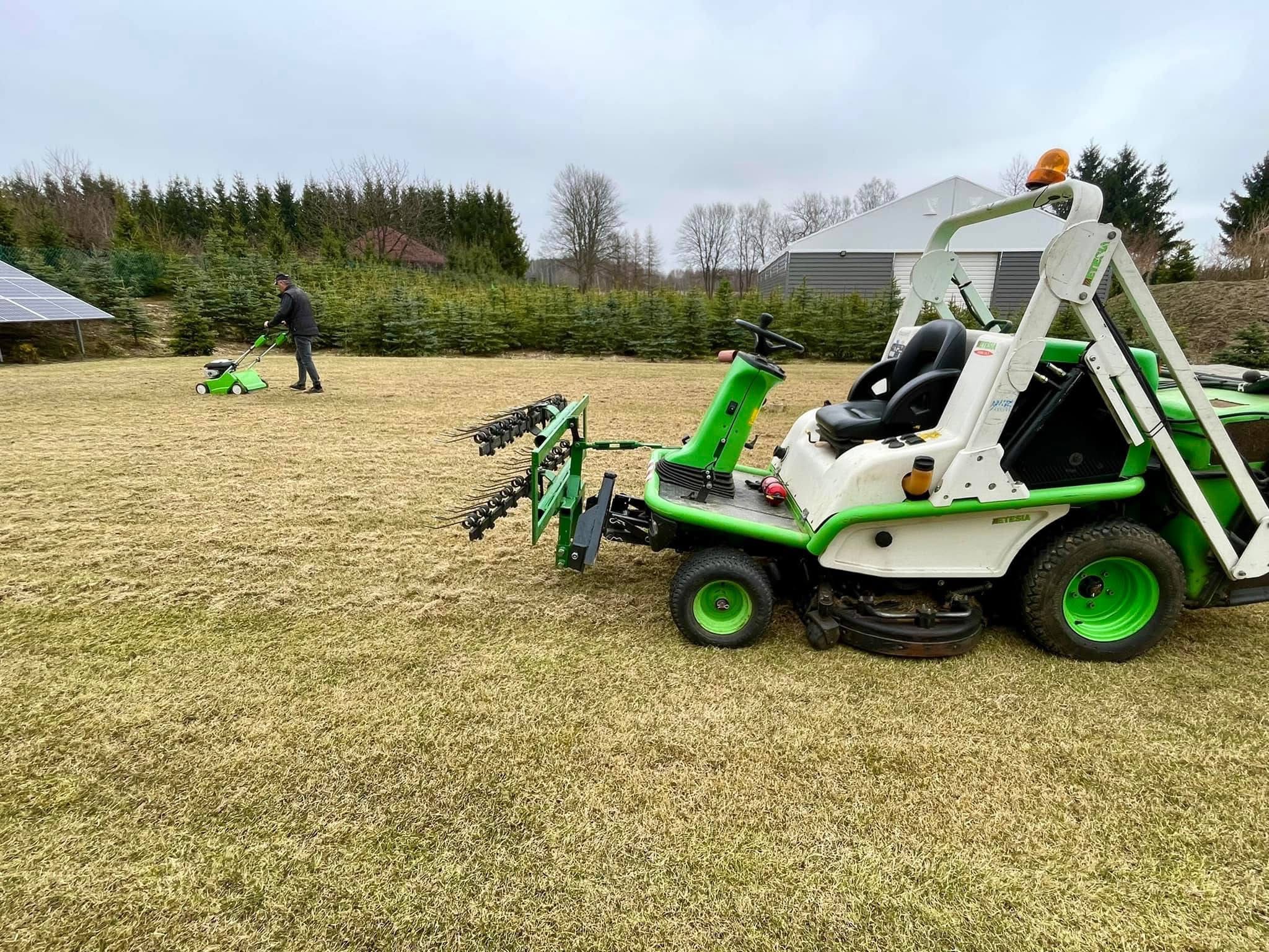 Profesjonalna kosiarka samojezdna z osprzętem do pielęgnacji trawnika na tle zieleni i budynku. W oddali osoba z kosiarką ręczną.