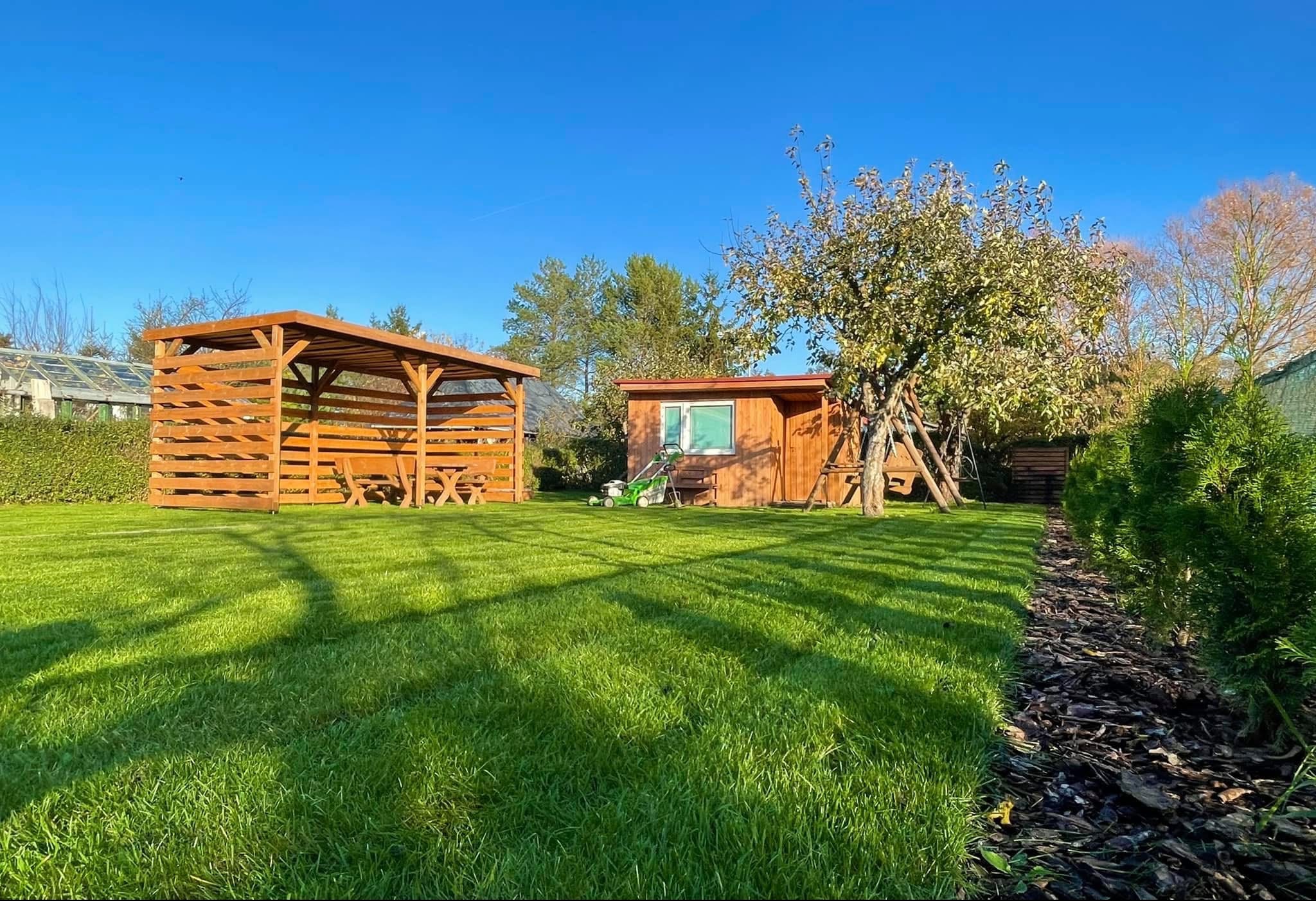 Zadbany ogród z równo skoszoną trawą, drewnianą altaną i domkiem narzędziowym w słoneczny dzień. Widoczna kosiarka i drzewo owocowe.