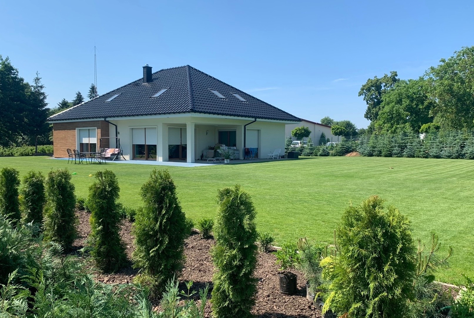 Zadbany ogród z równo przystrzyżonym trawnikiem i nasadzeniami iglaków przed nowoczesnym domem z ciemnym dachem i jasną elewacją w słoneczny dzień.