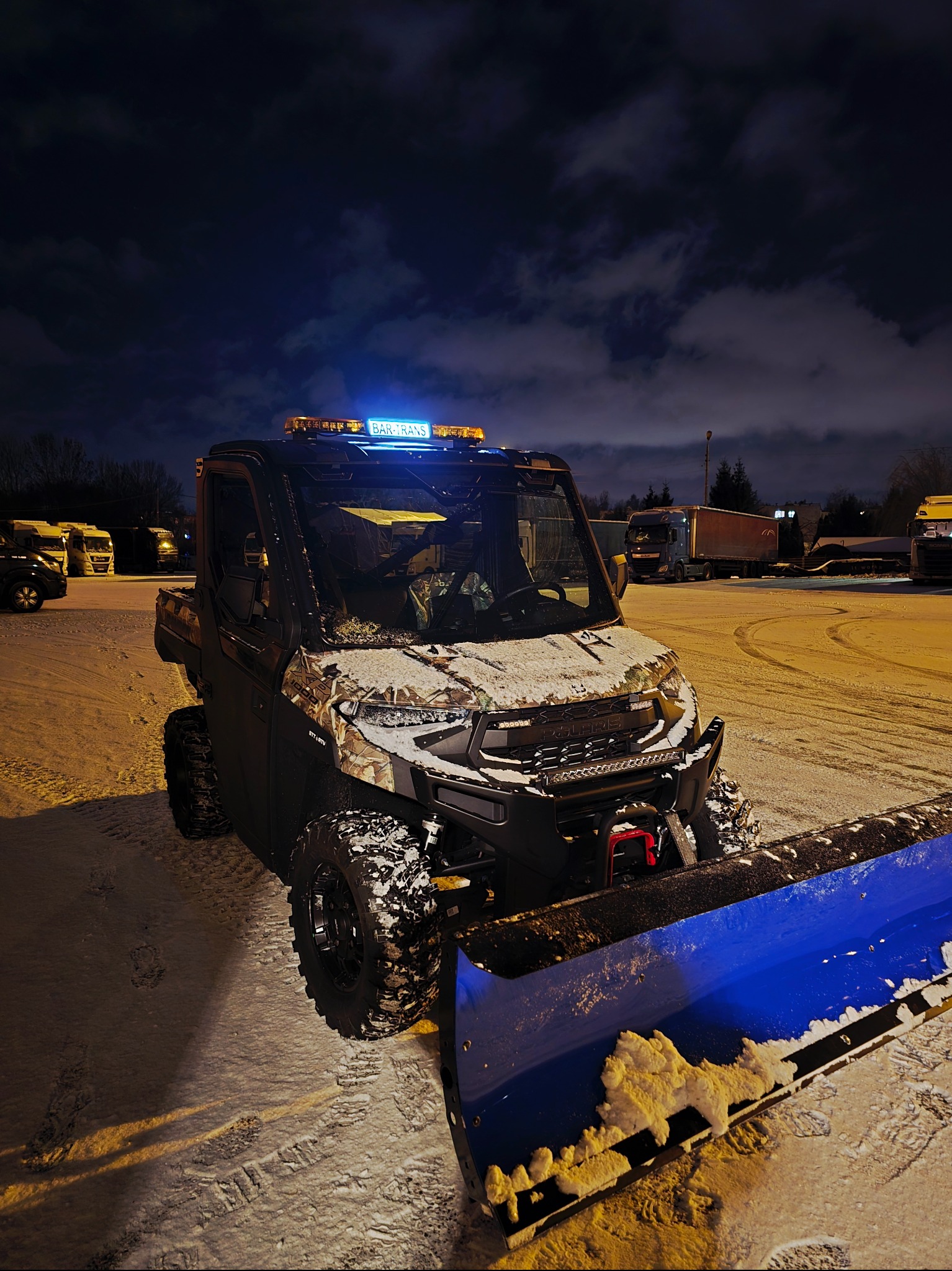 Pojazd z pługiem śnieżnym, oświetleniem ostrzegawczym i napisem 'BAR-TRANS' na dachu, zaparkowany na ośnieżonym placu w nocy, z widocznymi śladami opon.