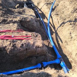 B&B Garden Usługi Ogrodnicze Hubert Rozynek - Instalacja nawadniania w wykopie: niebieskie rury złączone trójnikiem, czarna skrzynka zaworowa, narzędzia na ziemi. Etap prac w słoneczny dzień.