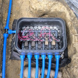 B&B Garden Usługi Ogrodnicze Hubert Rozynek - Instalacja systemu nawadniania w wykopie: skrzynka z zaworami, rury doprowadzające wodę. Widoczne kamienie i geowłóknina. Etap montażu.