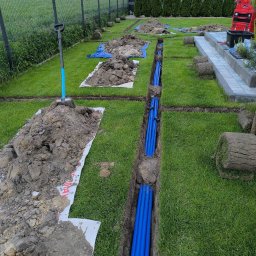 B&B Garden Usługi Ogrodnicze Hubert Rozynek - Instalacja niebieskich rur w wykopie na trawniku, przygotowanie pod system nawadniania. Widoczne rolki trawy i narzędzia ogrodnicze.
