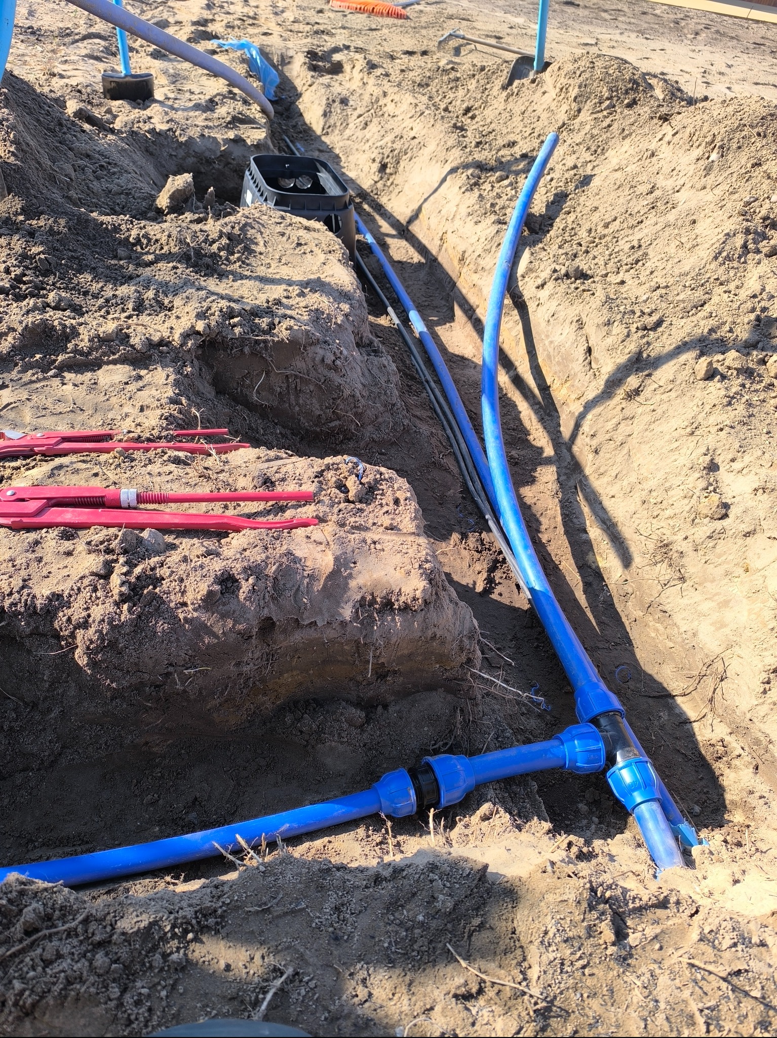 Instalacja nawadniania w wykopie: niebieskie rury złączone trójnikiem, czarna skrzynka zaworowa, narzędzia na ziemi. Etap prac w słoneczny dzień.