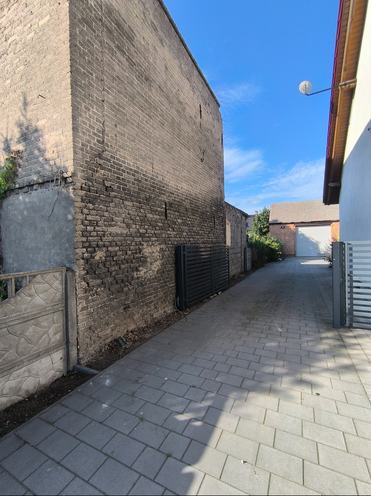 Ujęcie z poziomu kostki brukowej na posesji, ukazujące starą, ceglaną ścianę budynku z widocznymi śladami wilgoci i zanieczyszczeń, oraz fragment ogrodzenia.