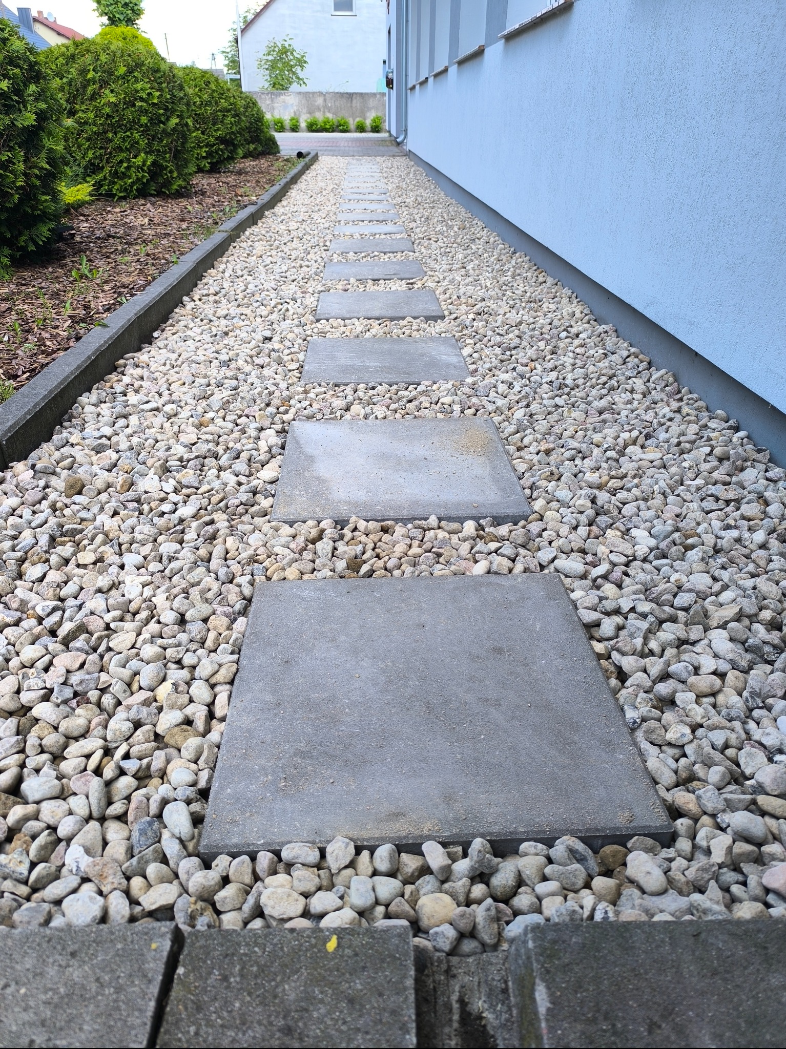 Ścieżka z betonowych płyt i jasnych kamieni prowadząca wzdłuż budynku, z widocznymi krzewami po lewej stronie. Minimalistyczny design ogrodu.