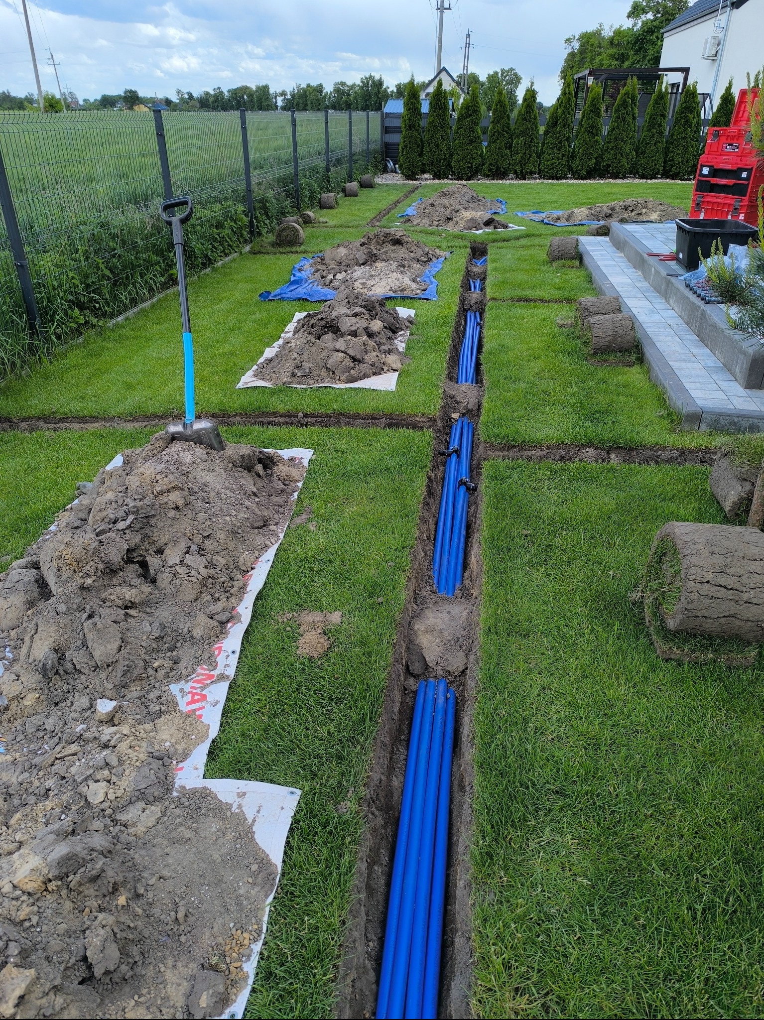 Instalacja niebieskich rur w wykopie na trawniku, przygotowanie pod system nawadniania. Widoczne rolki trawy i narzędzia ogrodnicze.