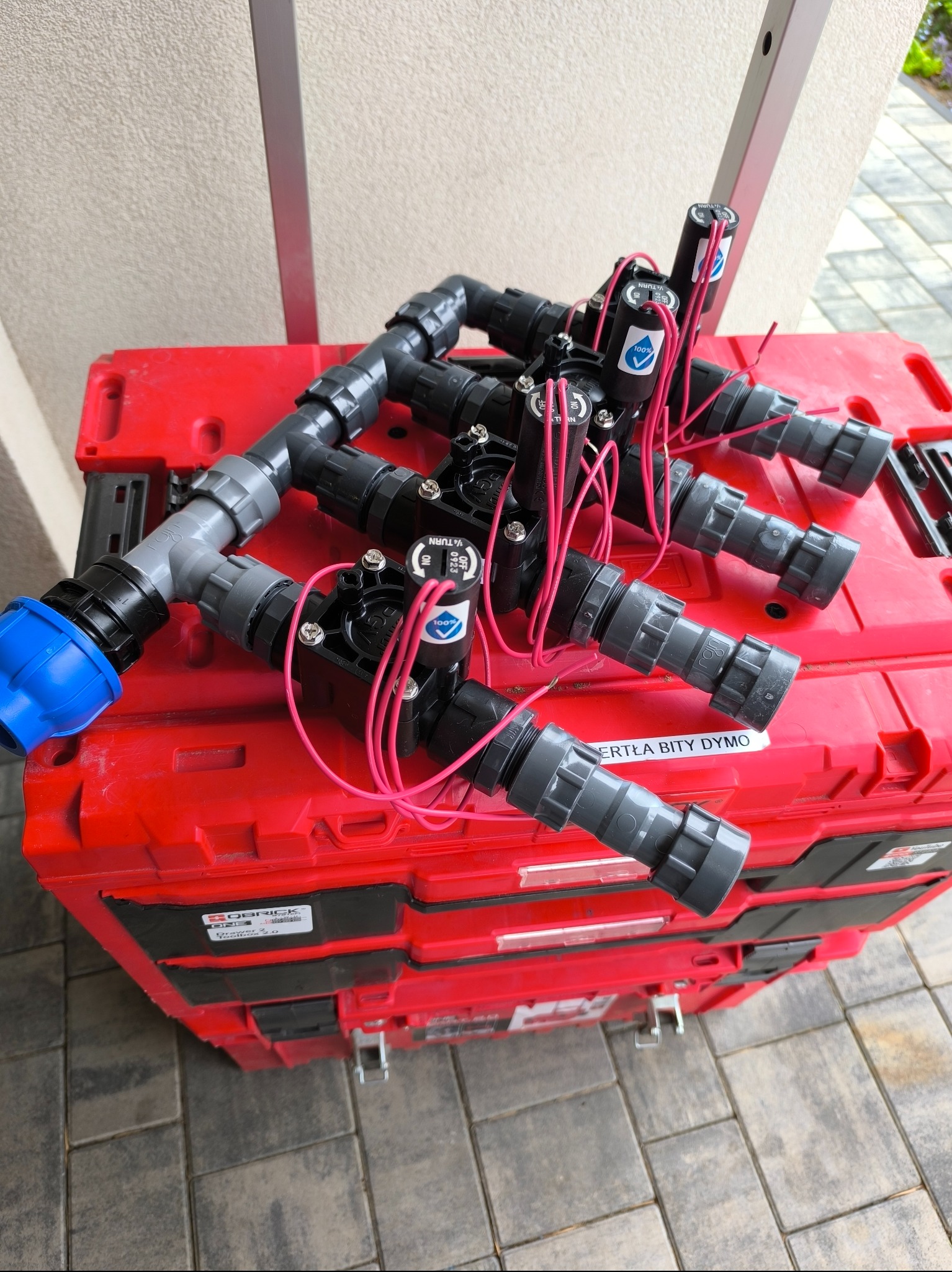Zestaw zmontowanych elektrozaworów do systemu nawadniania, z widocznymi przewodami i złączkami, ułożony na czerwonej skrzynce narzędziowej na zewnątrz.