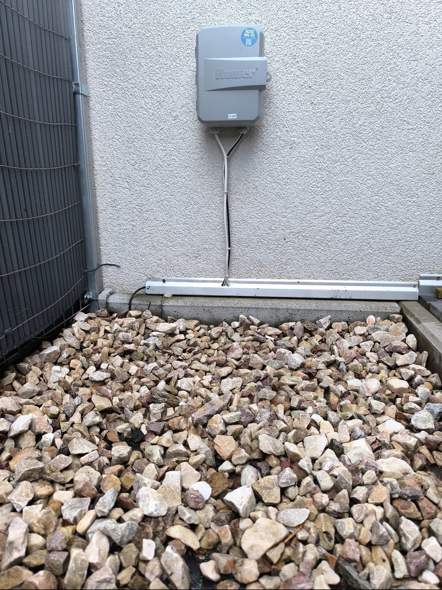Szary sterownik nawadniania Hunter na teksturowanej ścianie, z kablami i kamieniami dekoracyjnymi u podstawy. Widoczny fragment ogrodzenia.
