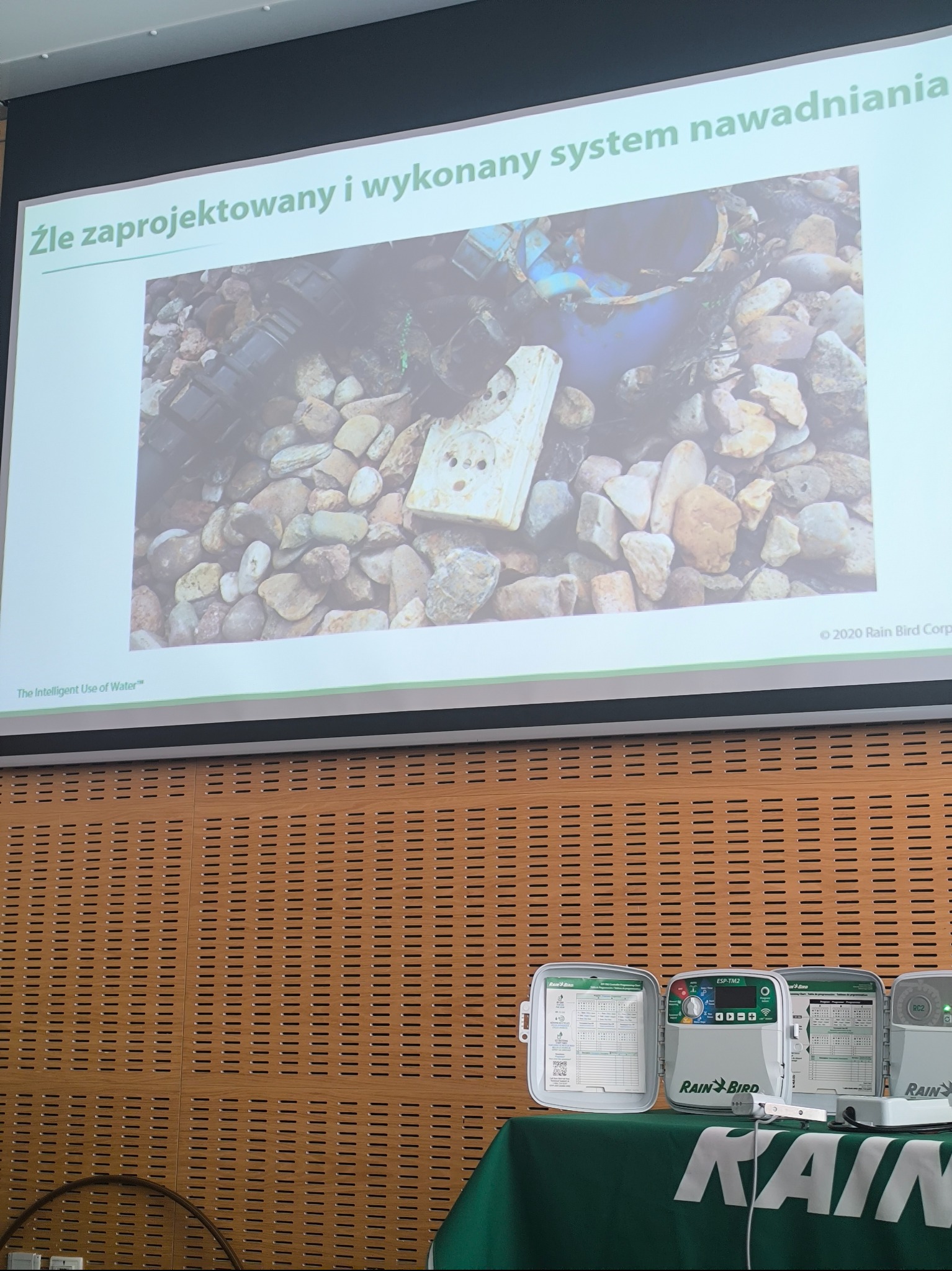 Prezentacja z przykładem źle zaprojektowanego systemu nawadniania, z widocznymi elementami instalacji na kamieniach i kontrolerami Rain Bird na stole.