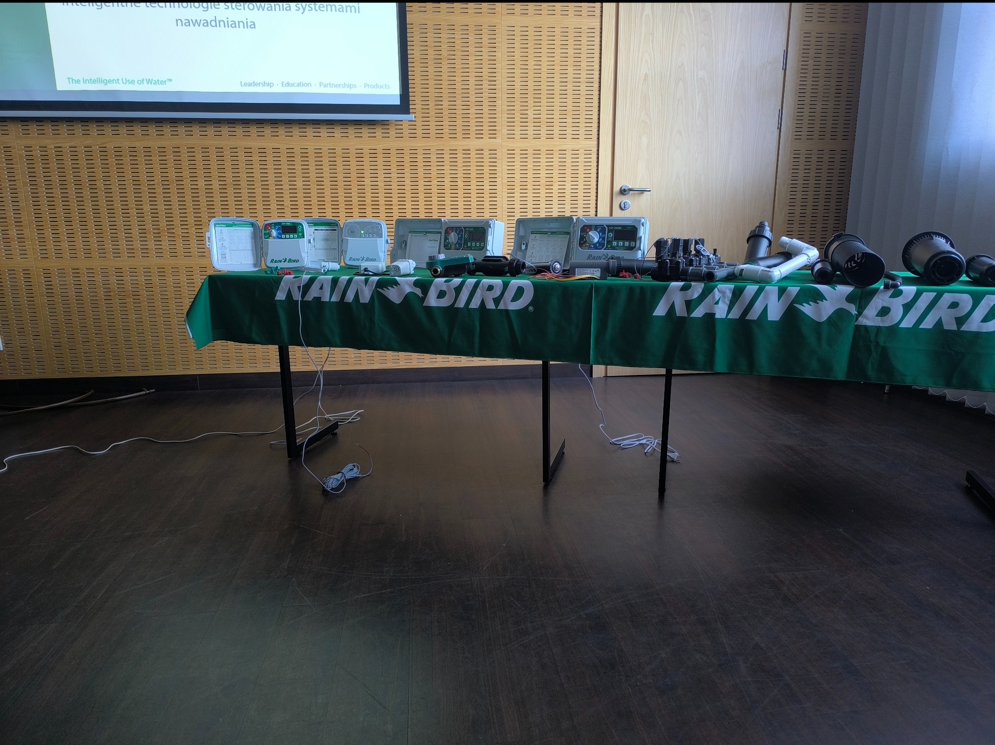 Prezentacja systemów nawadniania Rain Bird na stole z zielonym obrusem. Widoczne sterowniki, zraszacze i akcesoria. W tle ściana z panelami i ekran projekcyjny.