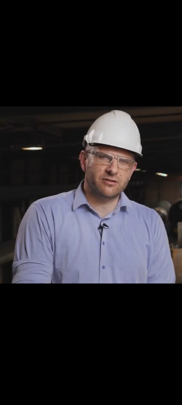 Inżynier w kasku i okularach, w jasnej koszuli, z mikrofonem krawatowym, na tle hali produkcyjnej z elementami konstrukcji stalowych. Ujęcie od pasa w górę.