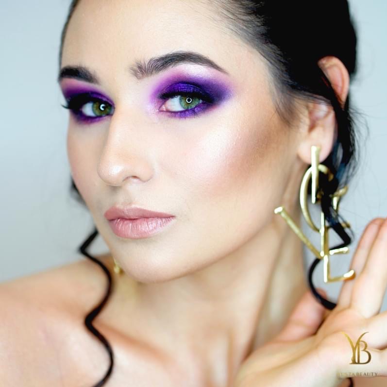 Portret kobiety z intensywnym fioletowym makijażem oczu, podkreślonymi kośćmi policzkowymi i dużymi złotymi kolczykami w kształcie logo YSL.