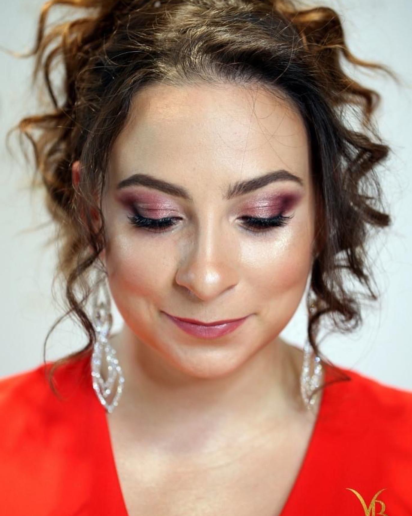 Portret kobiety z profesjonalnym makijażem oczu w odcieniach różu i fioletu, z błyszczącymi kolczykami i w czerwonej sukience, spoglądającej w dół.