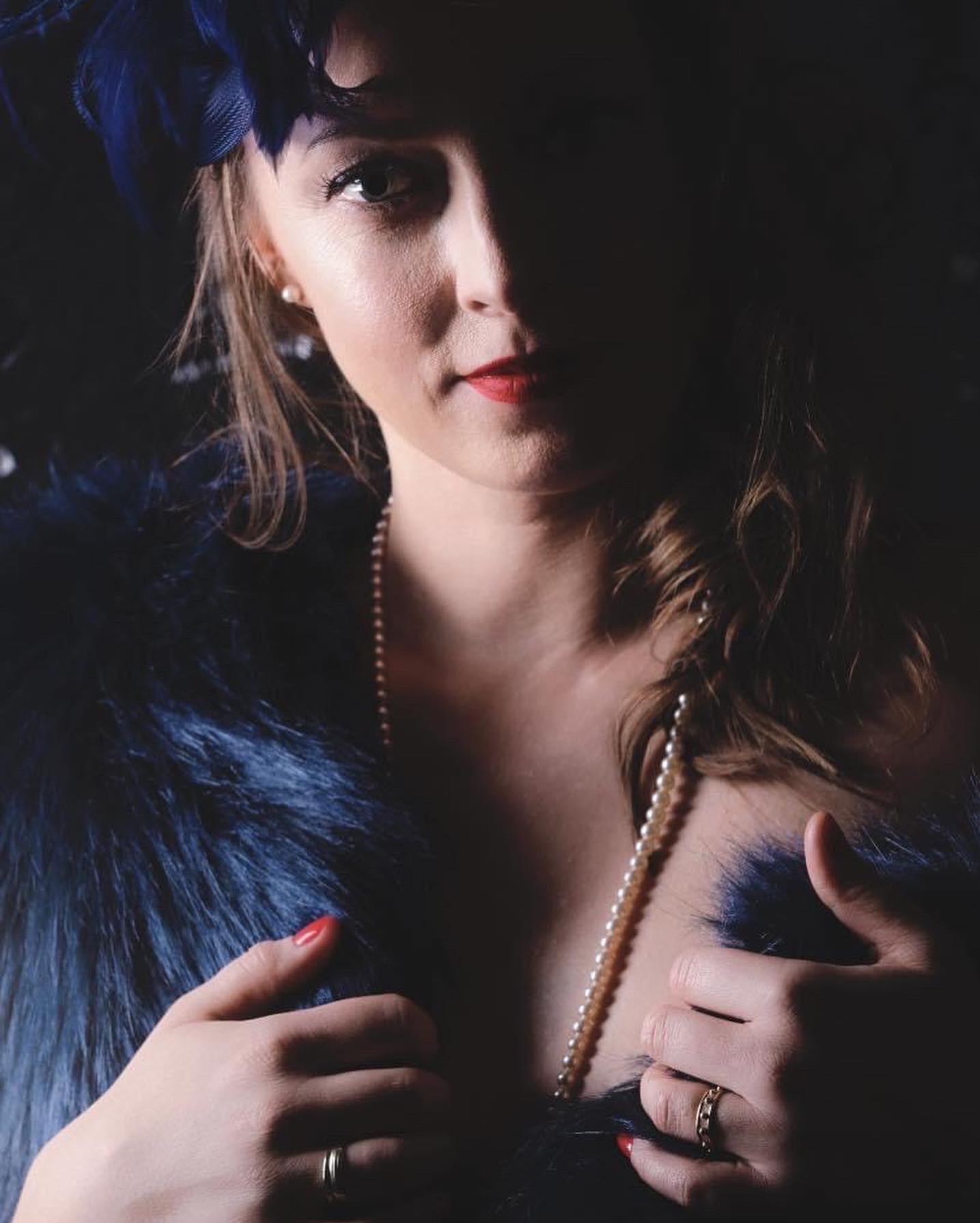 Portret kobiety w stylu retro z czerwonymi ustami, perłami i niebieskim boa. Część twarzy w cieniu, co dodaje dramatyzmu. Uwagę przyciąga pierścionek na palcu.