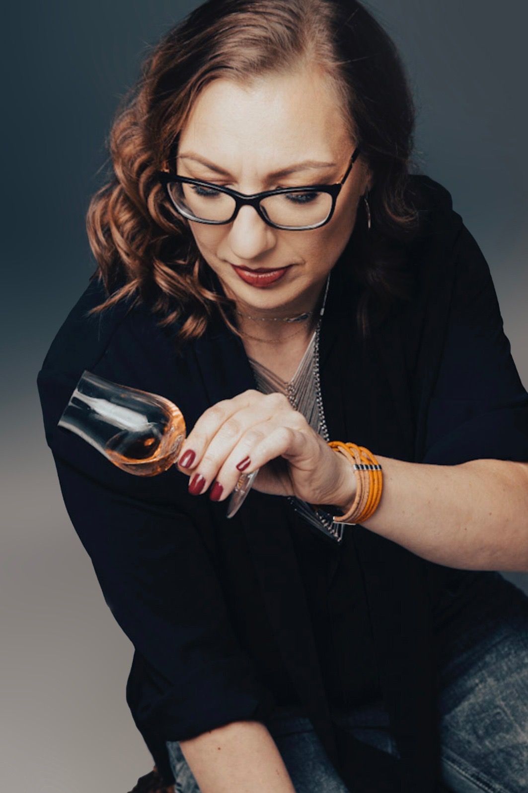 Kobieta w okularach, z czerwonymi ustami, trzyma kieliszek z bursztynowym płynem, ubrana w czarną marynarkę i dżinsy. Widoczne detale: manicure, biżuteria.