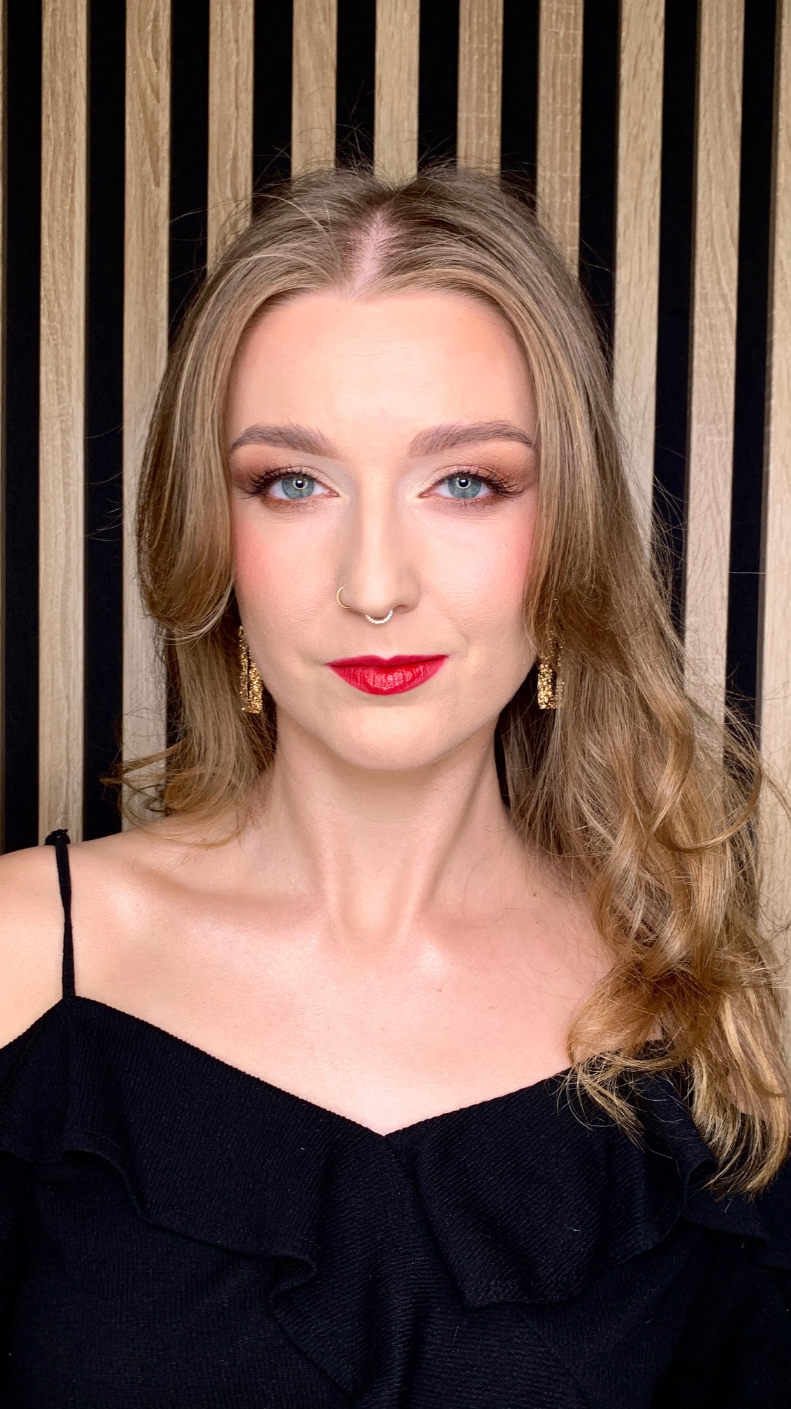 Portret kobiety z blond włosami, w czarnej sukience, z wyrazistym makijażem oczu i czerwonymi ustami, na tle ściany z drewnianych paneli.