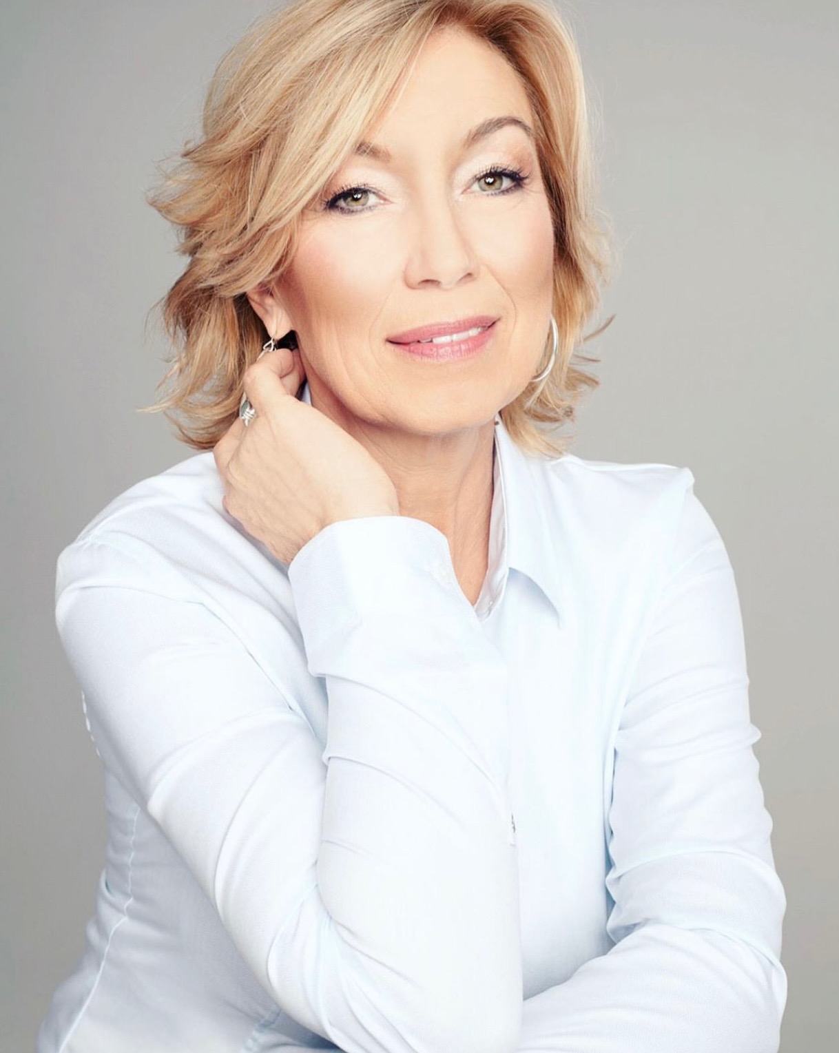 Portret kobiety w jasnej koszuli z profesjonalnym makijażem, delikatny uśmiech, blond włosy, srebrne kolczyki na szarym tle. Dłoń dotyka szyi.