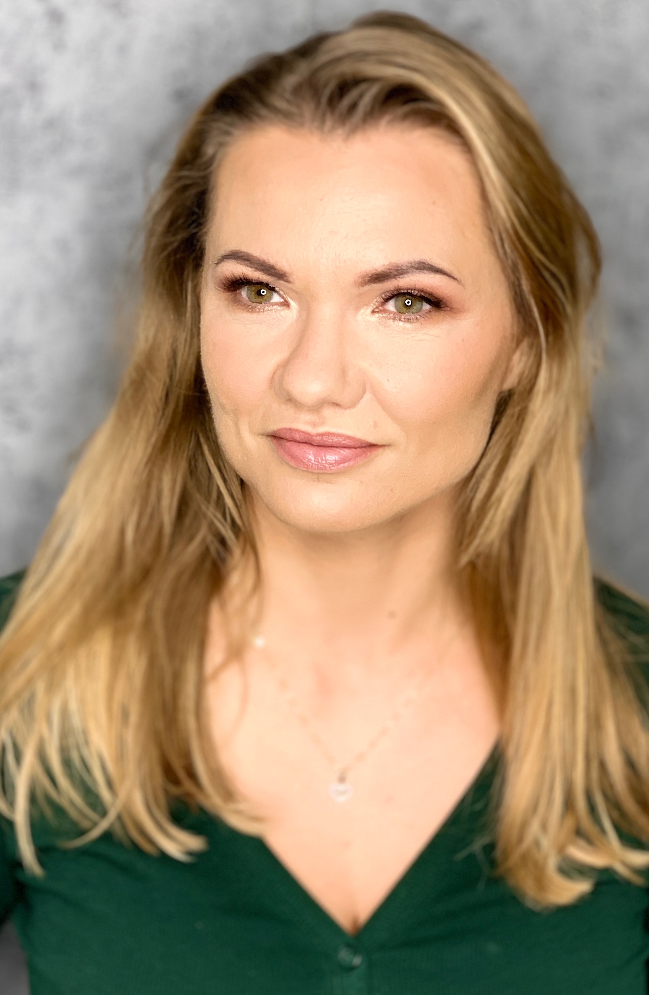 Portret kobiety z delikatnym makijażem w odcieniach brązu, podkreślającym zielone oczy. Blond włosy opadają na ramiona, a zielona bluzka dopełnia stonowaną kolorystykę.